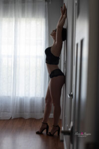A moody boudoir photo of a woman standing with her back against the wall and both arms are up above her head.